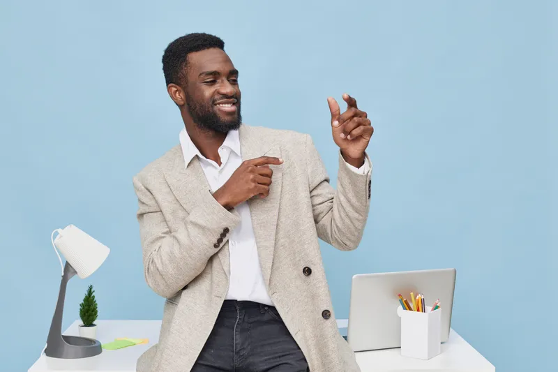 Smiling man wearing beige blazer and white shirt standing at desk with laptop, desk lamp, and stationery, cheerful expression, modern office background, lifestyle concept, professional, positive mood,