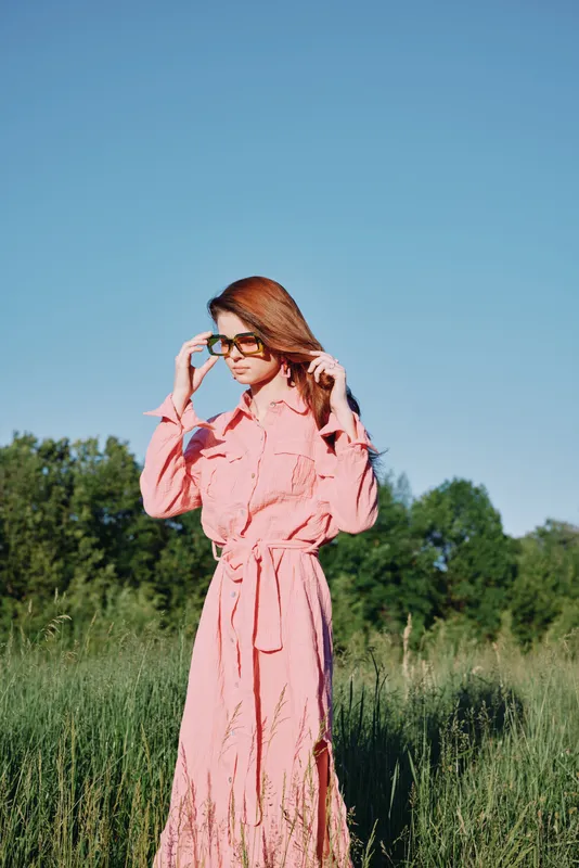 Young woman in a pink dress adjusting her sunglasses while standing in a field, showcasing summer fashion, relaxed mood, and a vibrant nature background