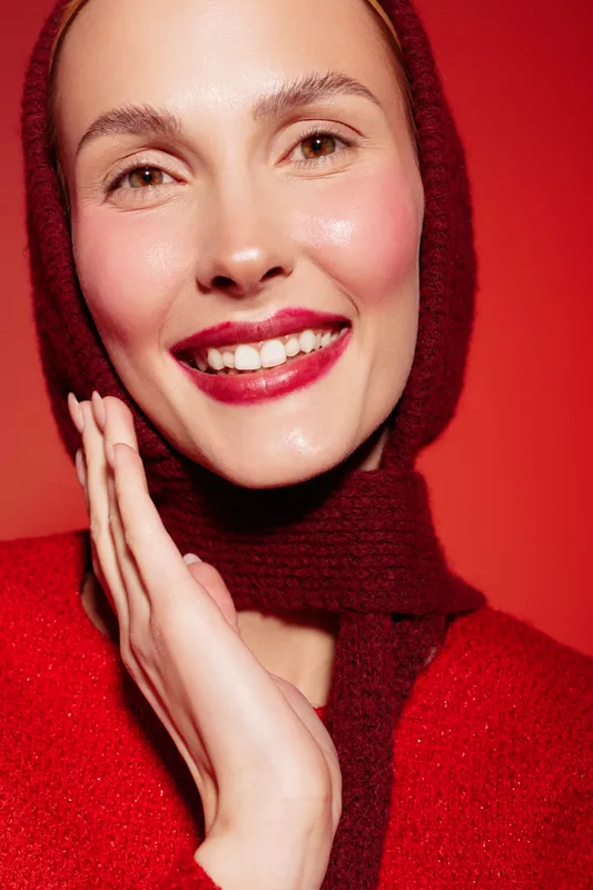 Smiling woman with red lips and scarf poses against red background. Close-up portrait shows natural skin glow and joyful expression. Concept of beauty, fashion, and happiness.