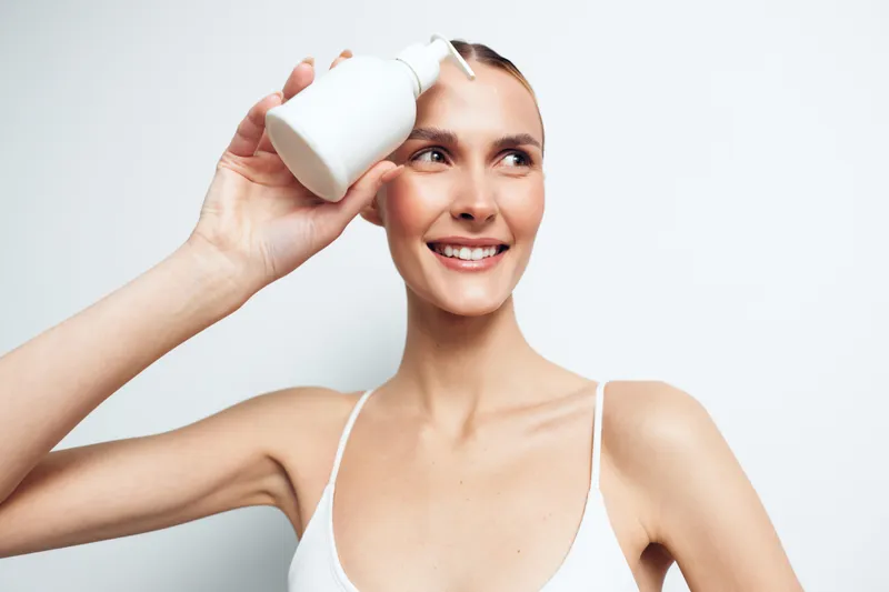 Smiling woman with clear skin holds white lotion bottle near face applying skincare product with positive expression isolated on white background in beauty concept.