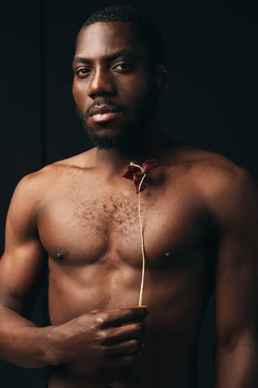 Black man with bare chest holding dried flower in creative conceptual visual style, portrait with serious expression and artistic mood on dark background.