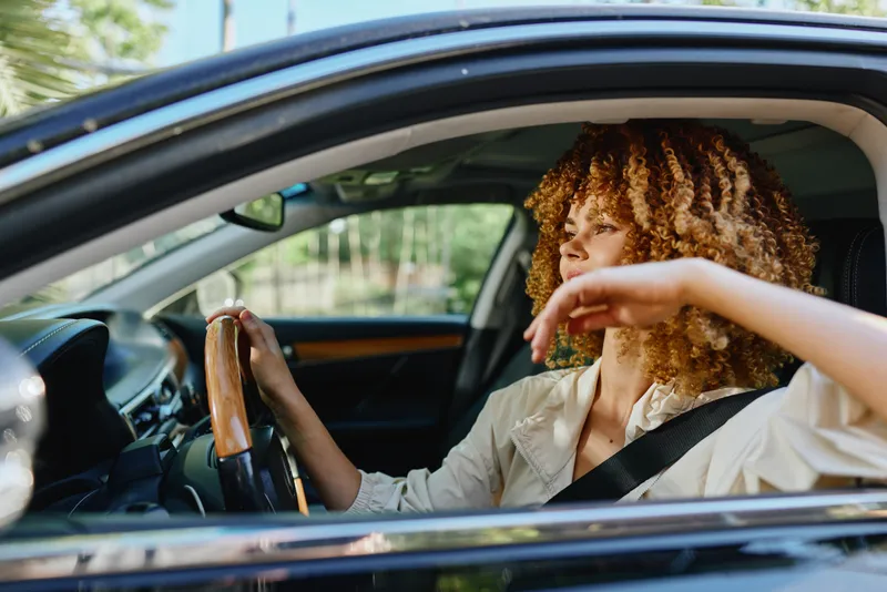 Young woman driving stylish black car with curly hair and beige jacket, enjoying sunny day with focus and confidence on the road outdoors.