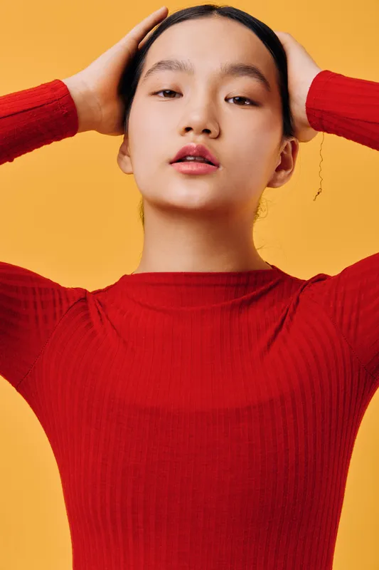 Young woman wearing red sweater with hands behind head posing confidently against orange background, looking straight at camera with natural expression and smooth skin.