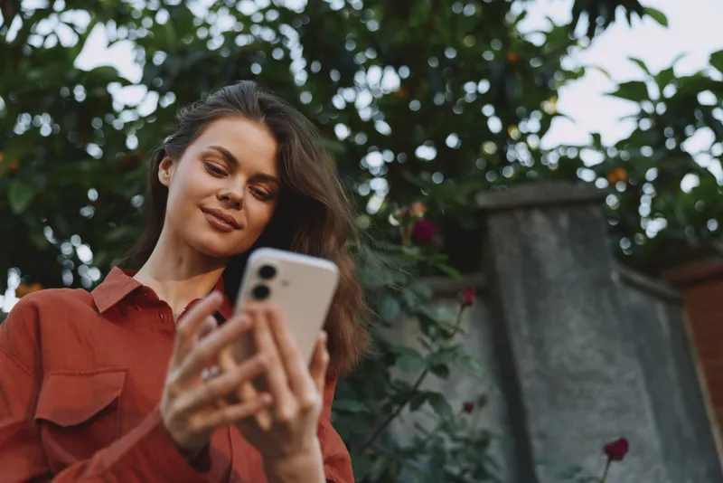 Cheerful woman outdoors, looking at smartphone, candid lifestyle moment, casual shirt, soft natural light, lush garden background, relaxed posture, warm tones, gentle breeze