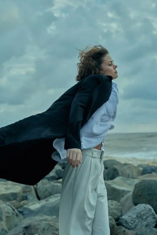 Wind, coat, ocean and sea set a dramatic scene of outdoor fashion, movement and windblown style, with a female figure in flowing clothing against rocks and a vast sky.