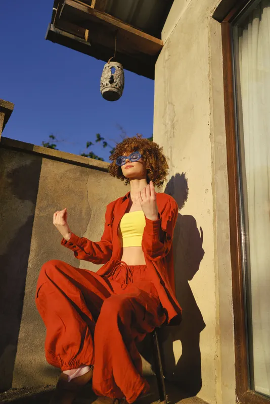 Energetic outdoor scene featuring a confident woman in a bright orange suit and yellow top, posing on a sunlit rooftop with relaxed lifestyle and bold fashion