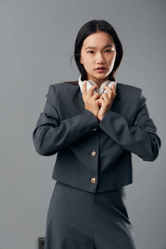 Young woman in gray formal suit holding white headphones close to chest with serious expression on neutral background, standing in studio for portrait or fashion shoot.