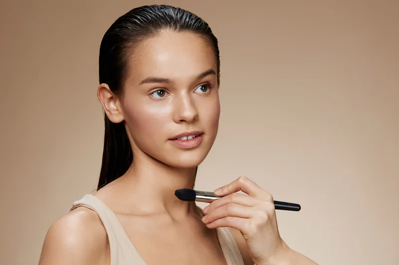 Young woman applying makeup with a brush, showcasing natural beauty and fresh skin tone, ideal for concepts of skincare, beauty rituals, and makeup artistry