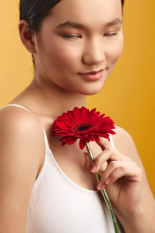 Asian woman gently holds a red flower close to her chest in a soft studio portrait, with a warm orange backdrop and a calm, contemplative mood.