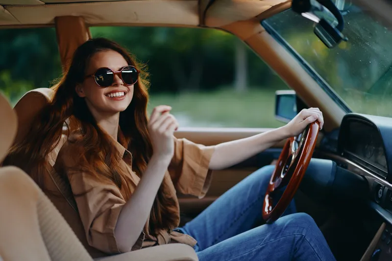 Happy young woman driving vintage car, wearing sunglasses, enjoying a sunny day, with lush green background, exuding joy and freedom.