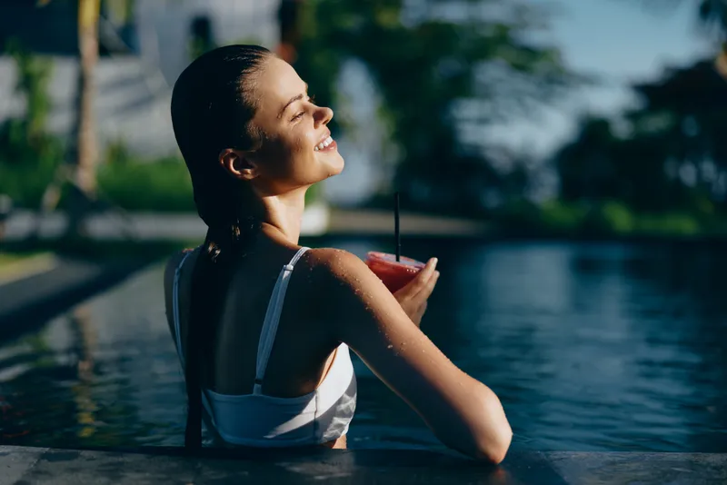 A relaxed woman lounges in a sunlit pool, sipping a pink beverage. Outdoor scene with water, palm trees, and bright daylight evokes leisure, happiness, and calm.