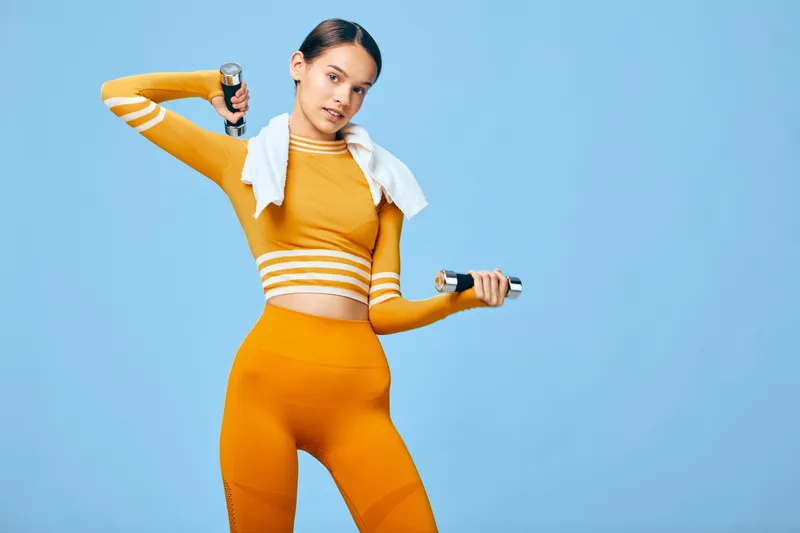 Female athlete in bright orange activewear lifts dumbbells in a dynamic workout. A white towel rests on her shoulders as she stands before a vibrant blue studio backdrop, showcasing athletic fitness