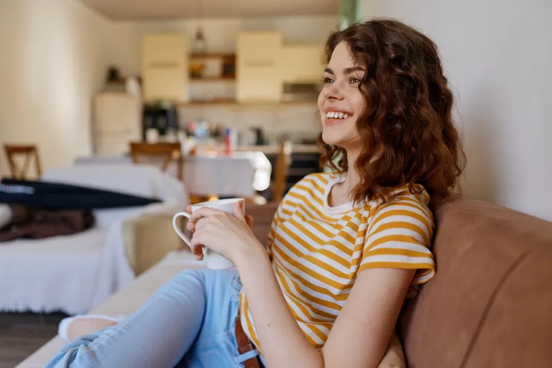 A woman relaxing at home on a sofa, wearing a striped t shirt, holding a mug and smiling, with soft daylight, cozy cushions, and a tidy modern living space.