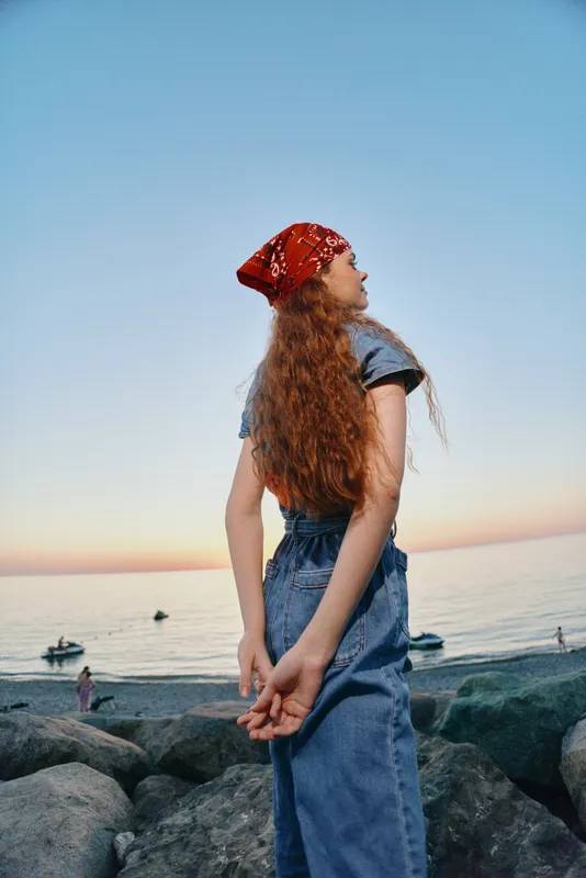 Lifestyle woman in modern boho-western denim street style with red bandana stands by seaside at sunset, film color capturing relaxed and natural mood.
