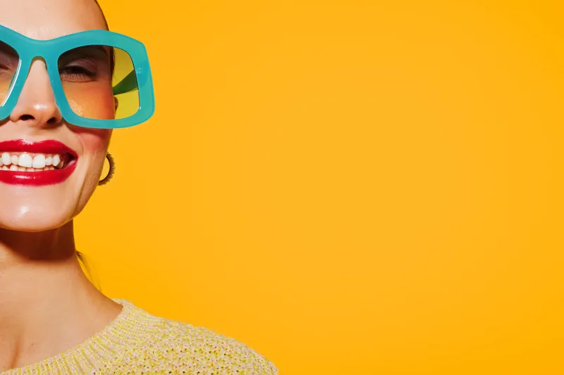 Close-up of smiling woman with bright red lipstick and large turquoise glasses on yellow background. Concept of fashion, beauty, summer style, and positive mood with vibrant colors.
