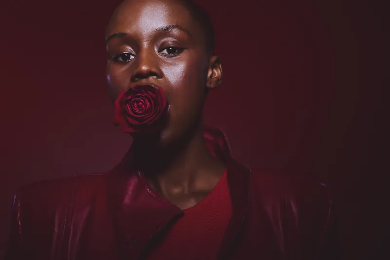 Portrait of a dark skinned African woman with a red rose in her mouth wearing a red leather jacket on a dark red background. Concept of beauty, fashion, and floral elegance.