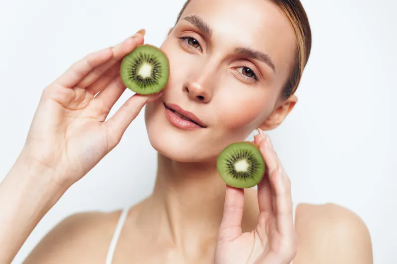 A young woman with clear skin holds two halves of a kiwi fruit near her face against a white background. The concept represents natural beauty and healthy lifestyle.