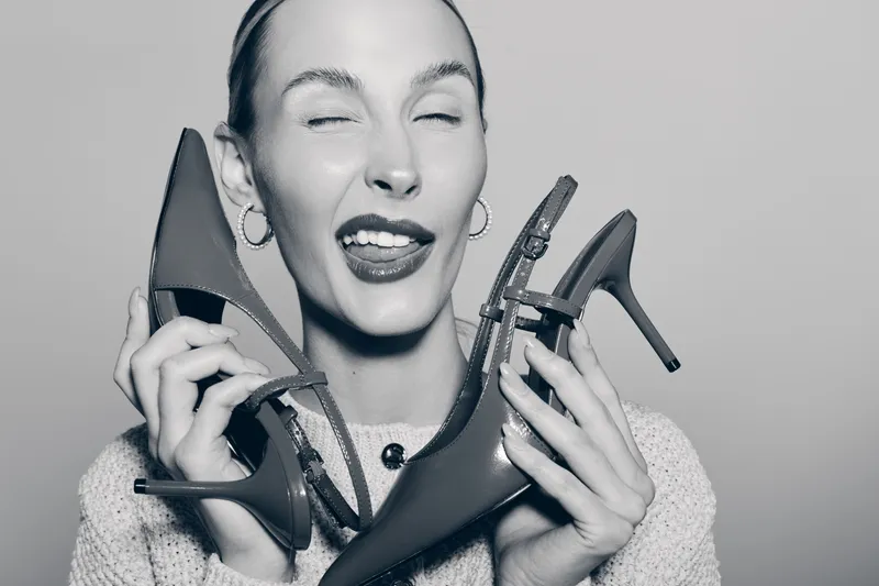 A young white woman with light skin holds red high heels near her face, eyes closed and playful expression in black and white photo.