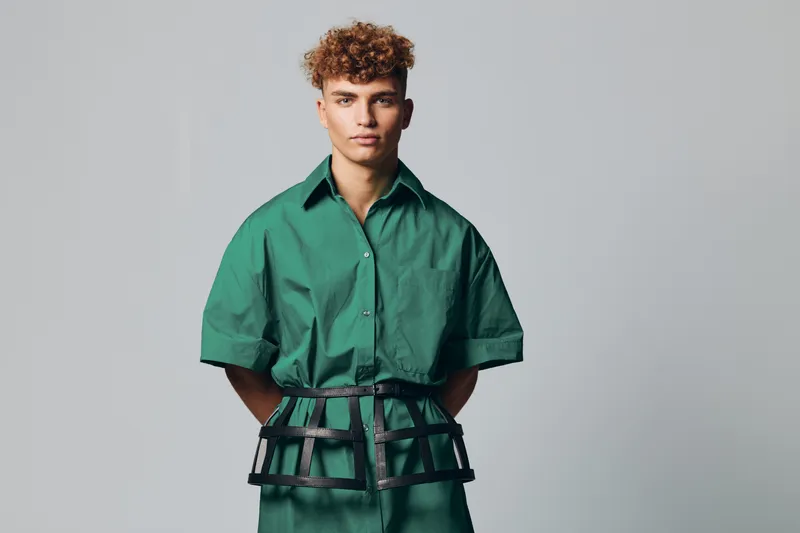 Young man with curly hair wearing green shirt and black harness posing against gray background in studio. Fashion model with serious expression and modern outfit for editorial shoot.