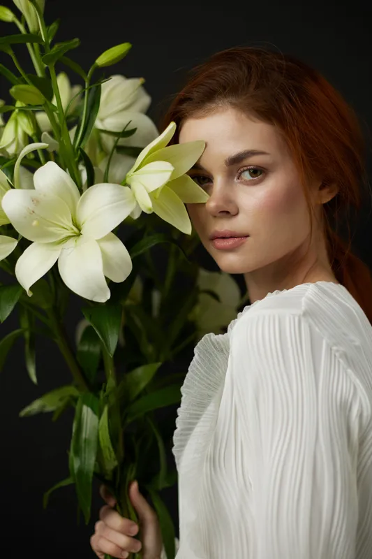 Beautiful young woman with brown hair holding white lilies on a dark background, expressing calm and elegance in a fashion forward, seasonal portrait
