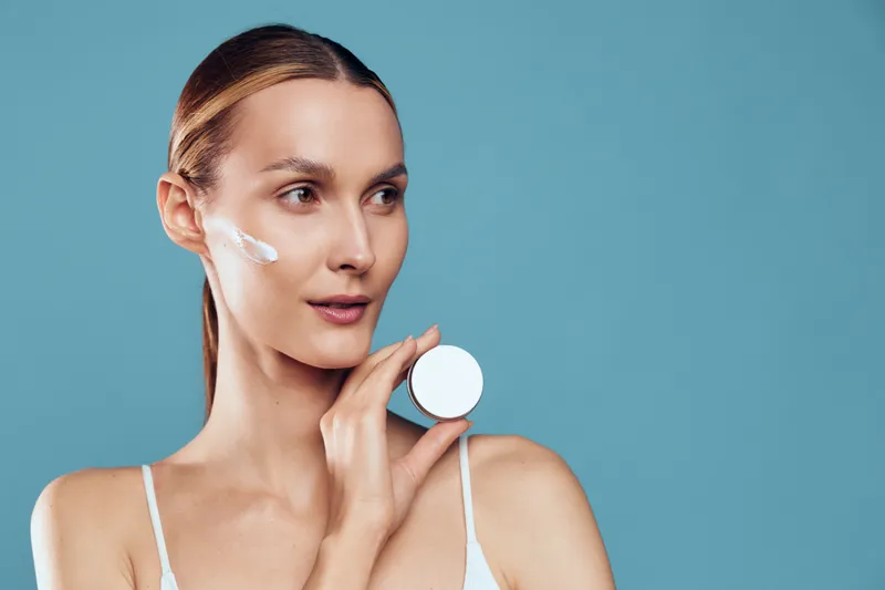 Bright portrait of a woman applying moisturizer to her cheek, cream on skin, calm expression, soft studio lighting, clean tones, skincare routine moment in focus, natural colors tones