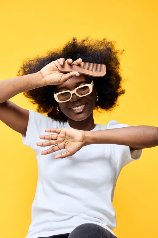 Happy young woman with natural afro hairstyle holding a comb, smiling playfully against a vibrant yellow background, embodying confidence and self care