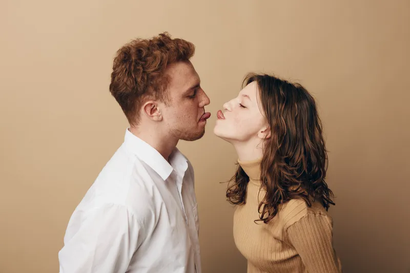 Young couple with curly hair and casual clothes leaning in for a kiss on beige background. Intimate moment, love, romance, and connection between man and woman in studio.