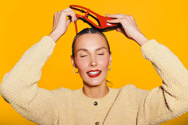 Young woman with closed eyes holds red high heel shoe above her head against bright yellow background. Concept of fashion, style, beauty, and playful mood in studio lighting.