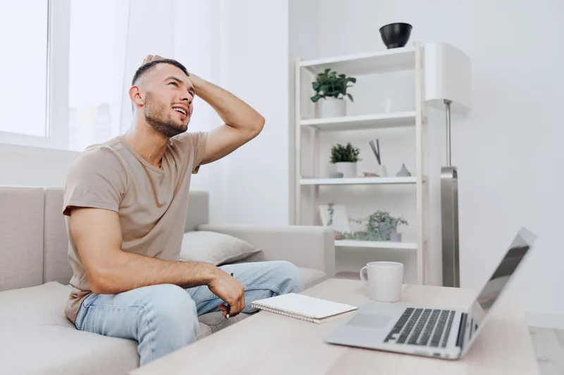 A stressed man sits on a couch, holding his head in frustration while working on a laptop. The calm background contrasts his emotions, illustrating modern work challenges.