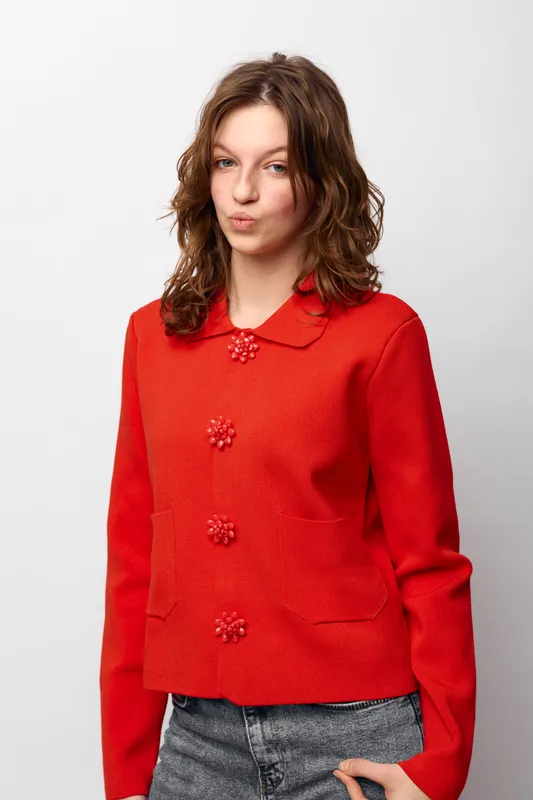 Young woman with curly brown hair wearing a red sweater with decorative buttons stands against white background. Casual style, confident expression, fashion, portrait, studio lighting.