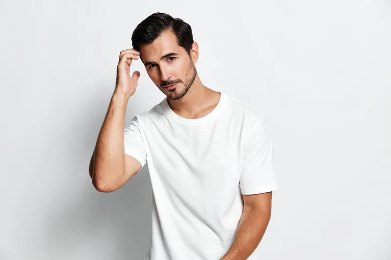 Attractive young man touching hair smiles confidently while posing in a casual white t-shirt against plain light background. Modern lifestyle concept.