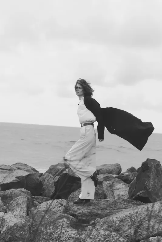 Woman stands on jagged rocks beside the sea, cape billowing in wind, grayscale fashion shot highlighting dramatic movement, ocean horizon, clothing elegance, natural coastal mood.
