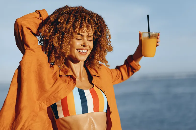 Cheerful woman outdoors by ocean, laughing in orange jacket with curly hair, holding a drink cup, sunny windy moment, soft light, relaxed confident pose, blue sea horizon today