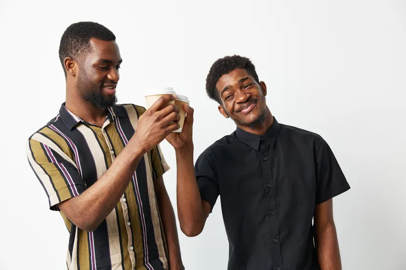 Two black men sharing a fun moment while clinking coffee cups, showcasing their friendship and joy. They are dressed casually and smiling together in a bright, modern atmosphere.