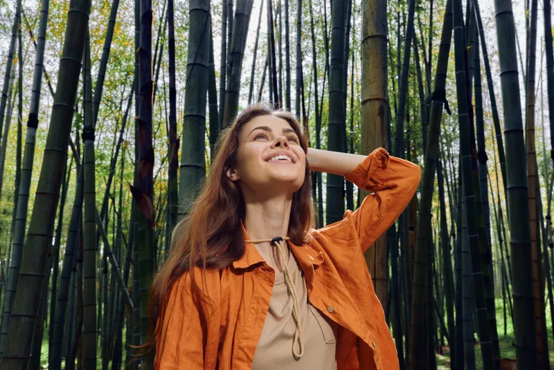 Woman in bamboo forest smiling and looking up, portrait of relaxed traveler in orange jacket, nature scene, outdoor happiness, peaceful mood and eco travel inspiration.