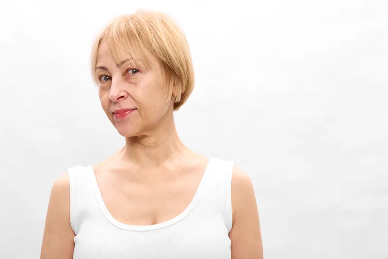 Mature woman with short blonde hair in white sleeveless top, studio portrait on white background, calm expression, concept of aging and beauty care daily.