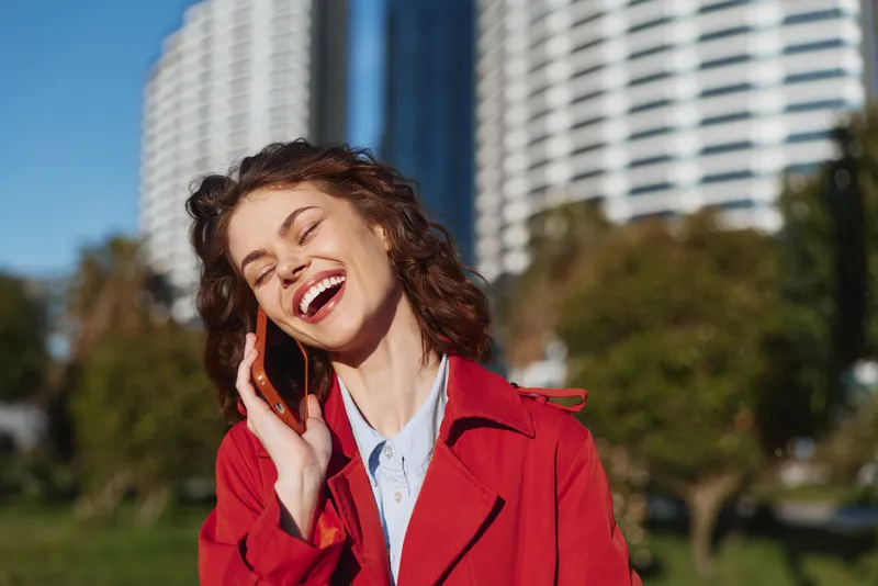 Cheerful woman outdoors, smiling with eyes closed, sunlit moment, relaxed pose, soft breeze, bold red coat, urban park backdrop, green grass, modern buildings nearby, clear blue sky