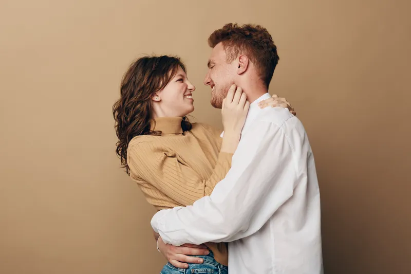 Smiling young couple embraces and looks at each other with affection on beige background. Concept of love, relationship, happiness, and intimacy between partners.