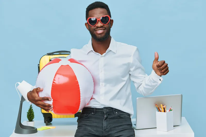 Vibrant smiling man with dark skin and afro hairstyle wearing stylish white shirt and heart-shaped sunglasses holding a colorful inflatable beach ball at a modern office workspace with laptop and stat