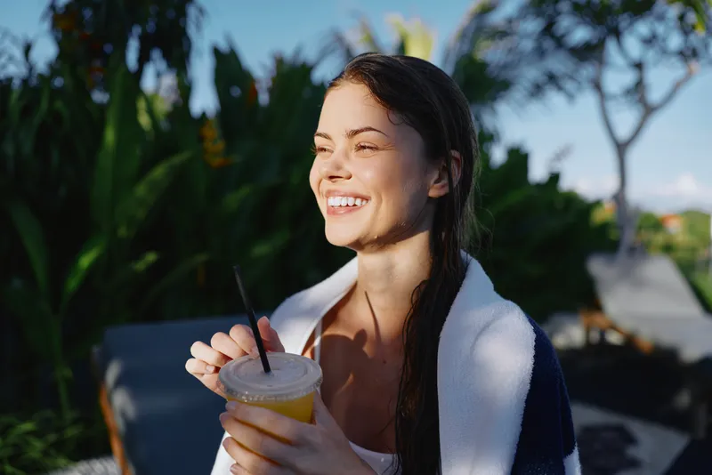 Cheerful woman outdoors, smiling while holding a refreshing drink, sunlit moment, relaxed posture, natural colors, lush greenery in background, casual summer lifestyle scene