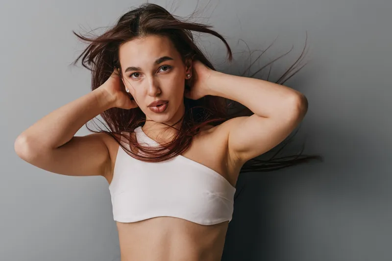 Young woman with long hair posing against gray background, wearing white crop top, hands behind head, looking at camera with confident expression and natural makeup in studio.