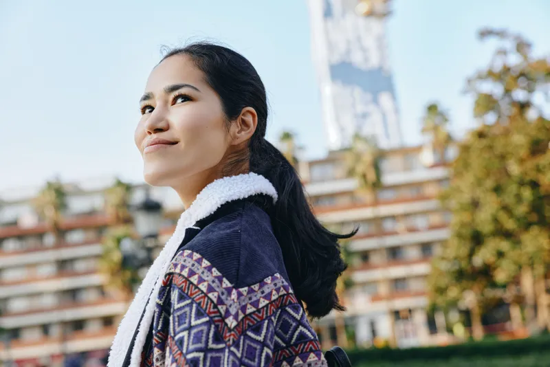 Woman outdoors in an urban setting, wearing a patterned jacket and scarf, looking up with a hopeful smile, city background, seasonal fashion mood, candid lifestyle.