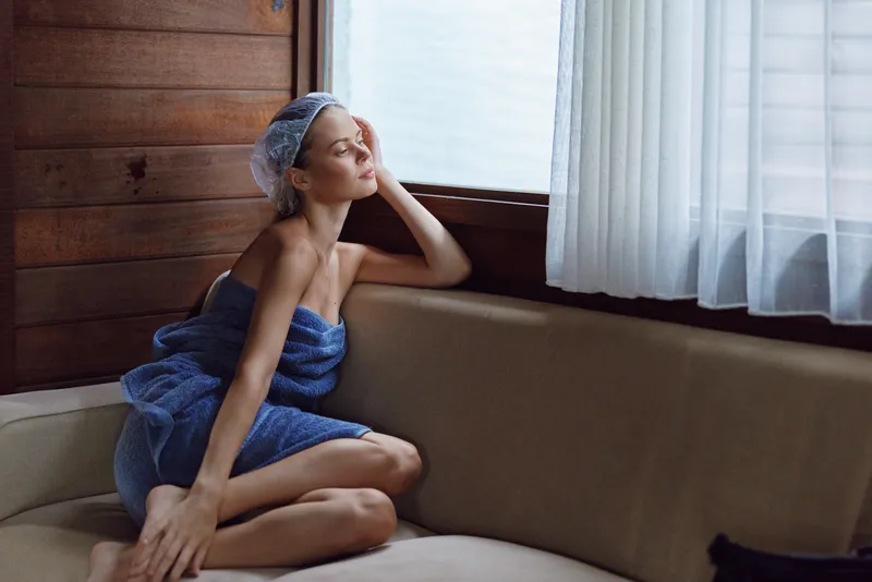 Relaxed woman wrapped in a blue towel and hair cap sitting on a cozy beige sofa indoors near wooden walls and soft natural light from a window with white curtains.