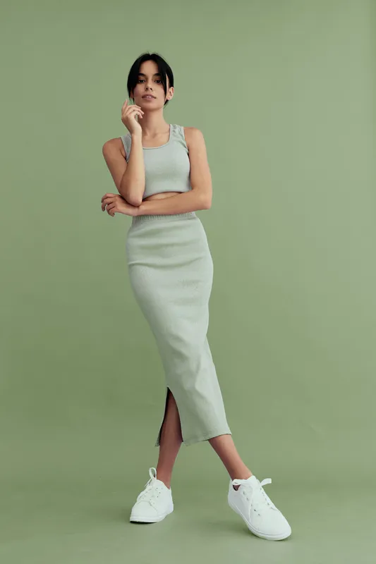 young woman standing confidently in a light green dress and white sneakers against a plain green background, casual style and relaxed pose
