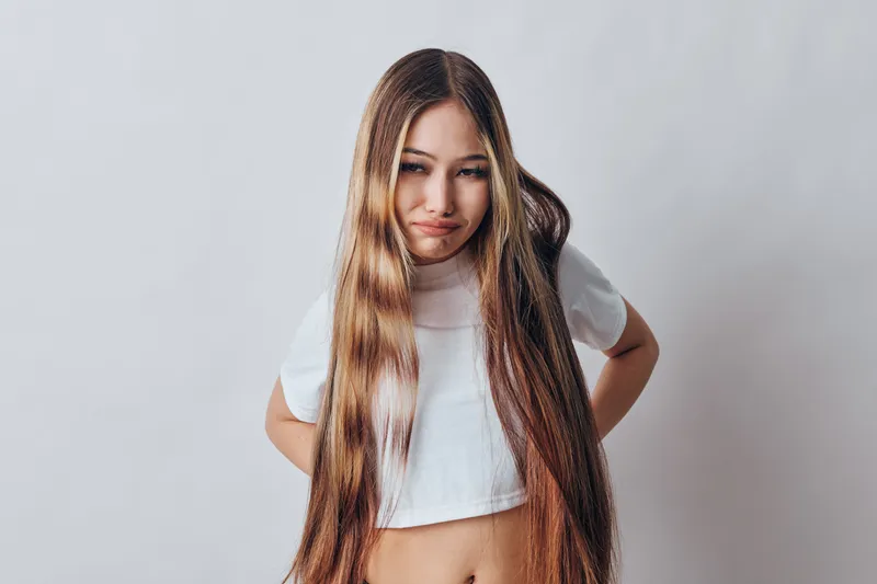 Young girl with long straight hair wearing white crop top stands against plain background with hands on hips looking at camera in confident casual pose for portrait.