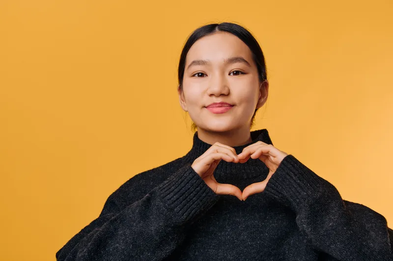 Young woman with black sweater making heart shape with hands smiling at camera against orange background. Portrait of female expressing love and positive emotion with gesture.