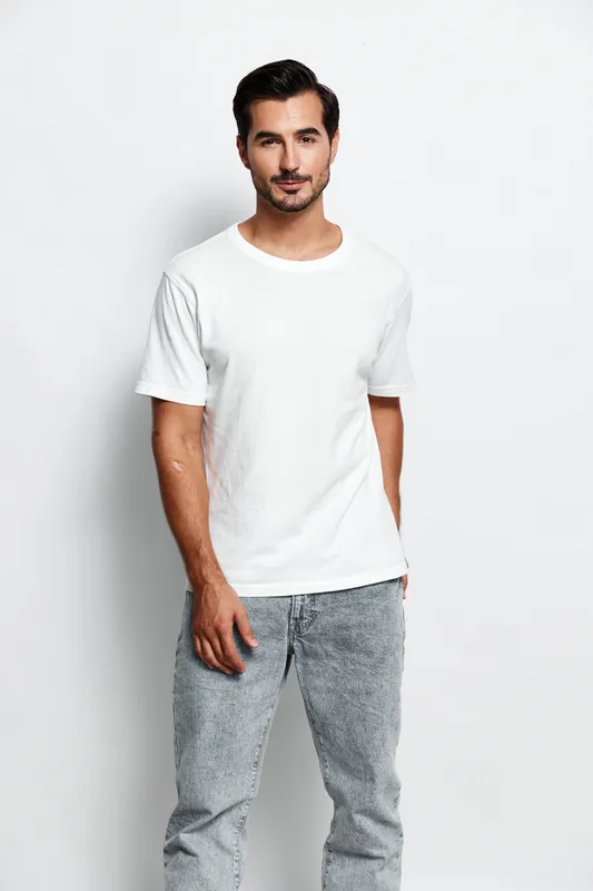 Full body confident young man dressed in casual white t-shirt and grey jeans stands smiling against a solid light background, showcasing a relaxed, modern style.