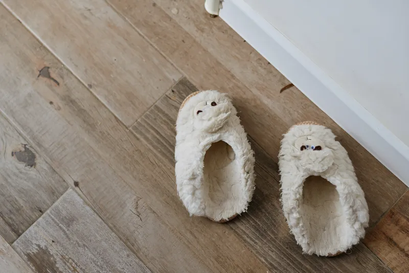 Cozy fluffy slippers rest on a wooden floor beside a white wall, capturing a warm intimate home lifestyle moment with soft textures, quiet comfort and everyday coziness.
