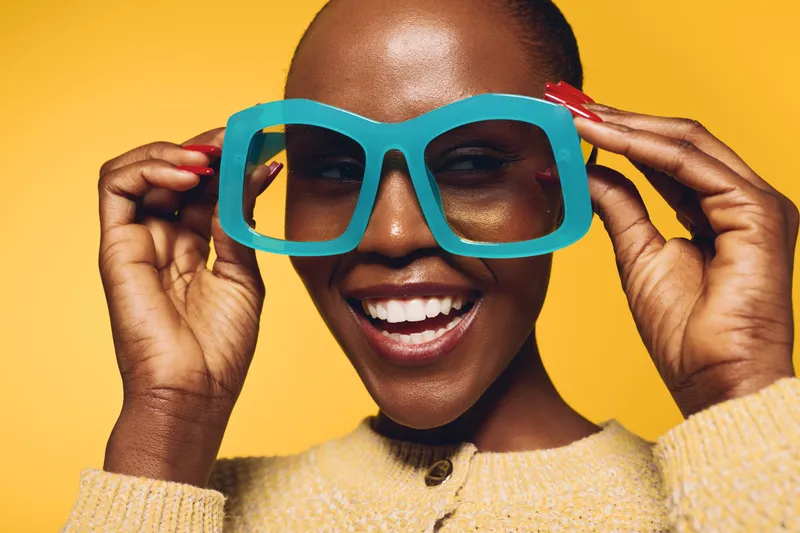 Smiling dark skinned African woman with short hair wearing large blue glasses and yellow sweater on yellow background. Concept of fashion, beauty, happiness, and confident lifestyle.