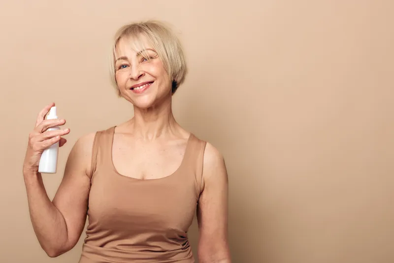 Smiling mature woman with short blonde hair sprays facial mist on skin, wearing beige tank top against neutral background. Concept for skincare, hydration, self-care, and beauty routine.
